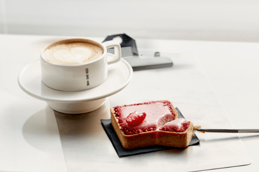 Zdjęcie przedstawia filiżankę z kawą. Obok leży prostokątne ciastko z malinową polewą.