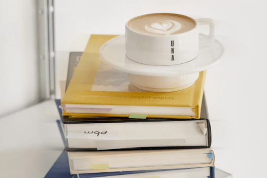 The photo shows a pile of books with a cup of coffee standing on top.